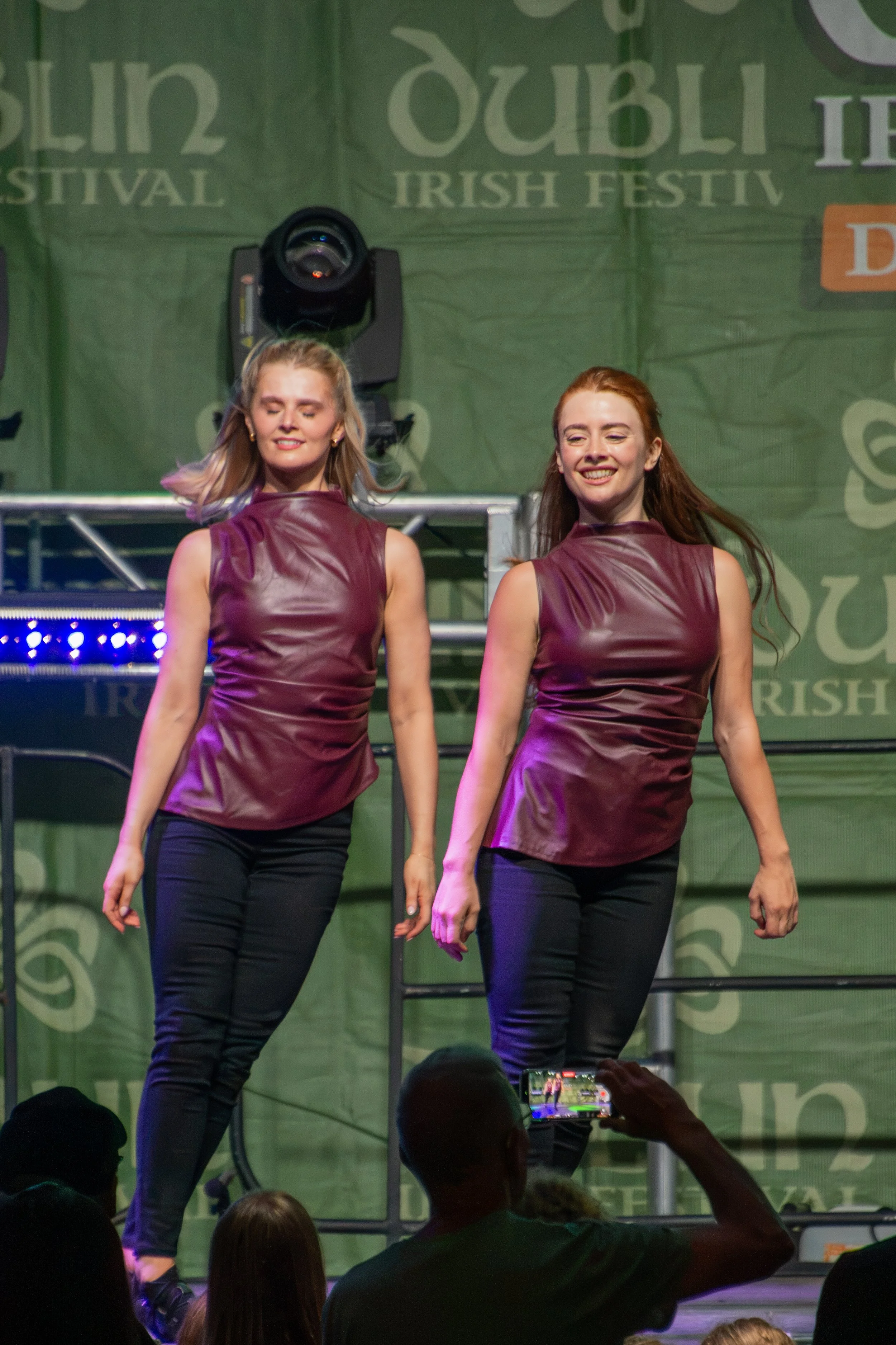 Two women in maroon sleeveless tops and black pants dancing on stage at a St. Patrick's Festival event, with a green background displaying text and large stage lights behind them, and an audience watching and recording the performance.