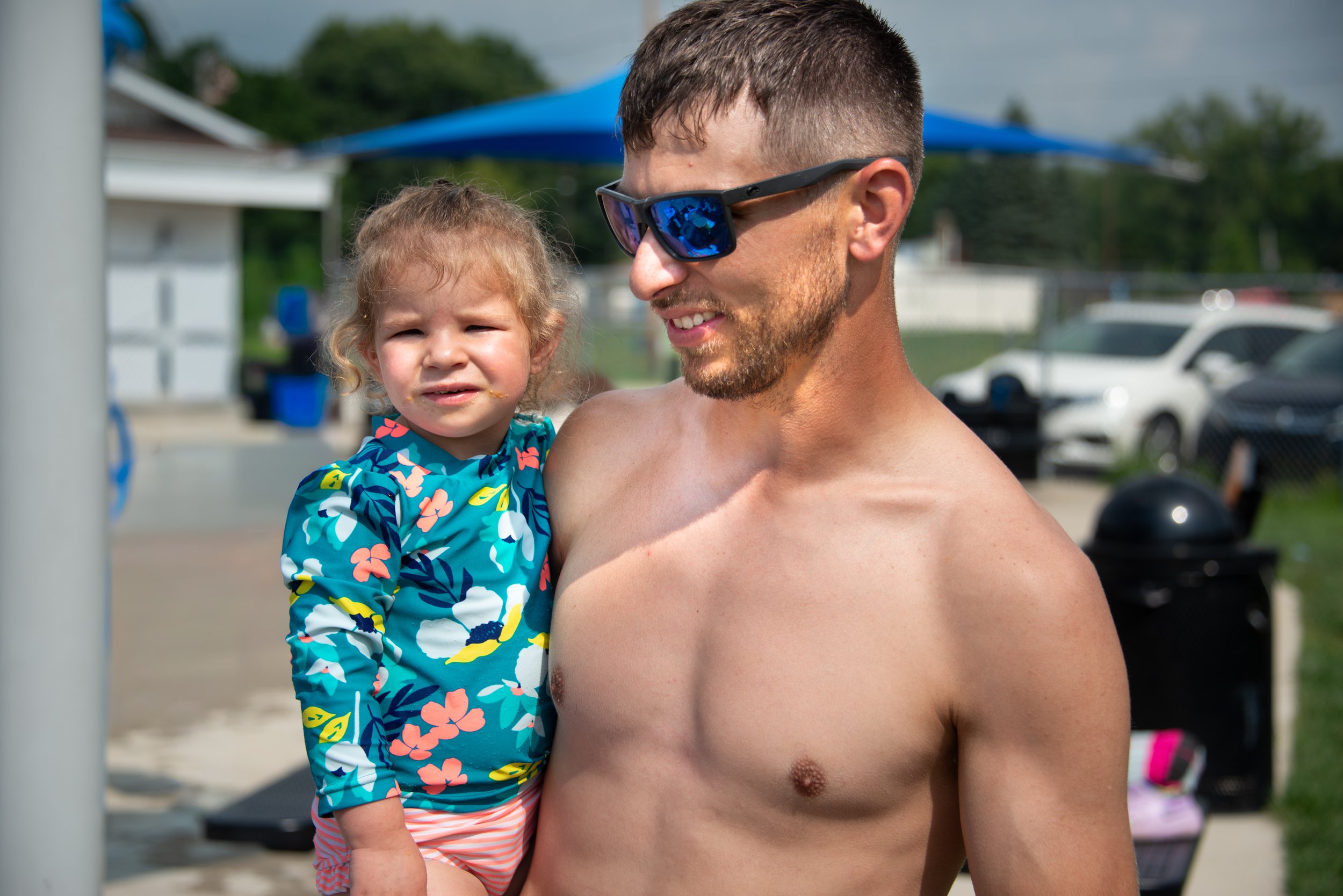 A shirtless man wearing sunglasses holding a young girl in a colorful swimsuit at a poolside or outdoor water park.