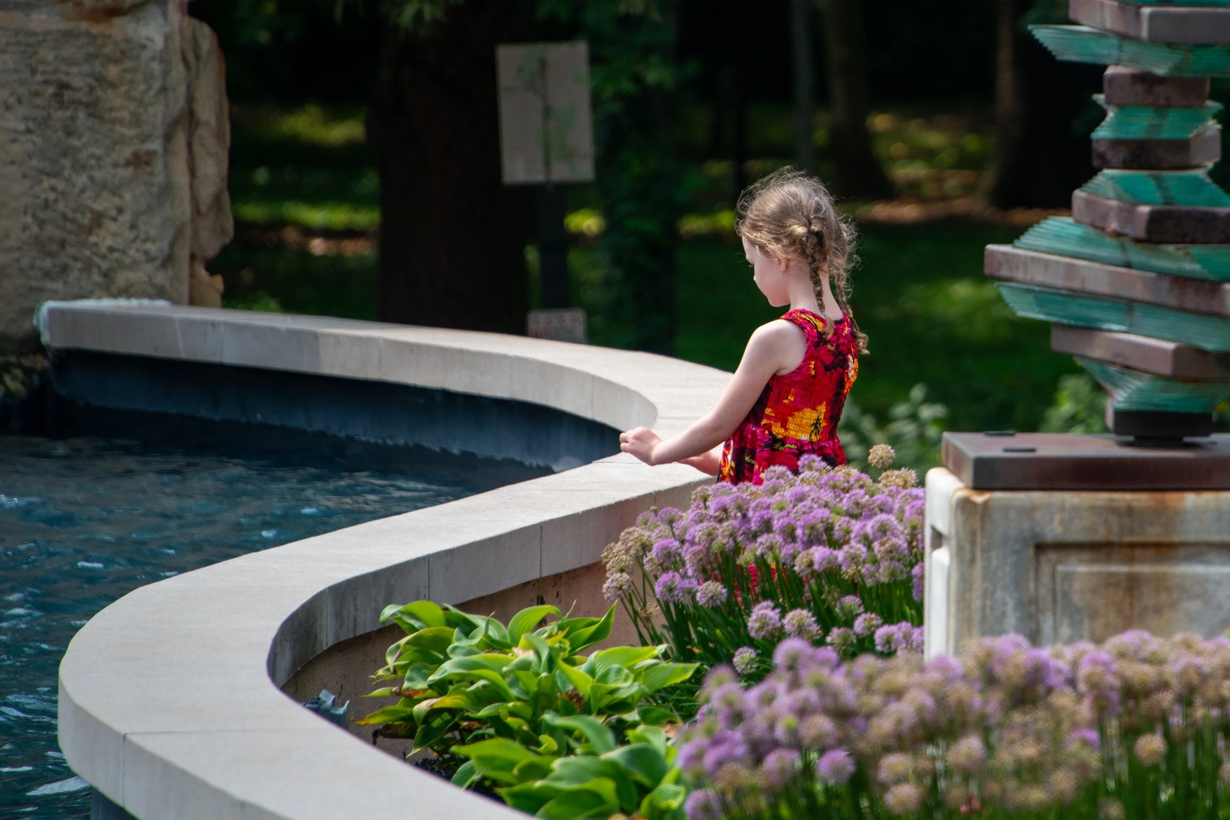 A young girl with braided hair and a colorful dress stands by a curved stone water fountain in a garden, surrounded by pink flowers and greenery.