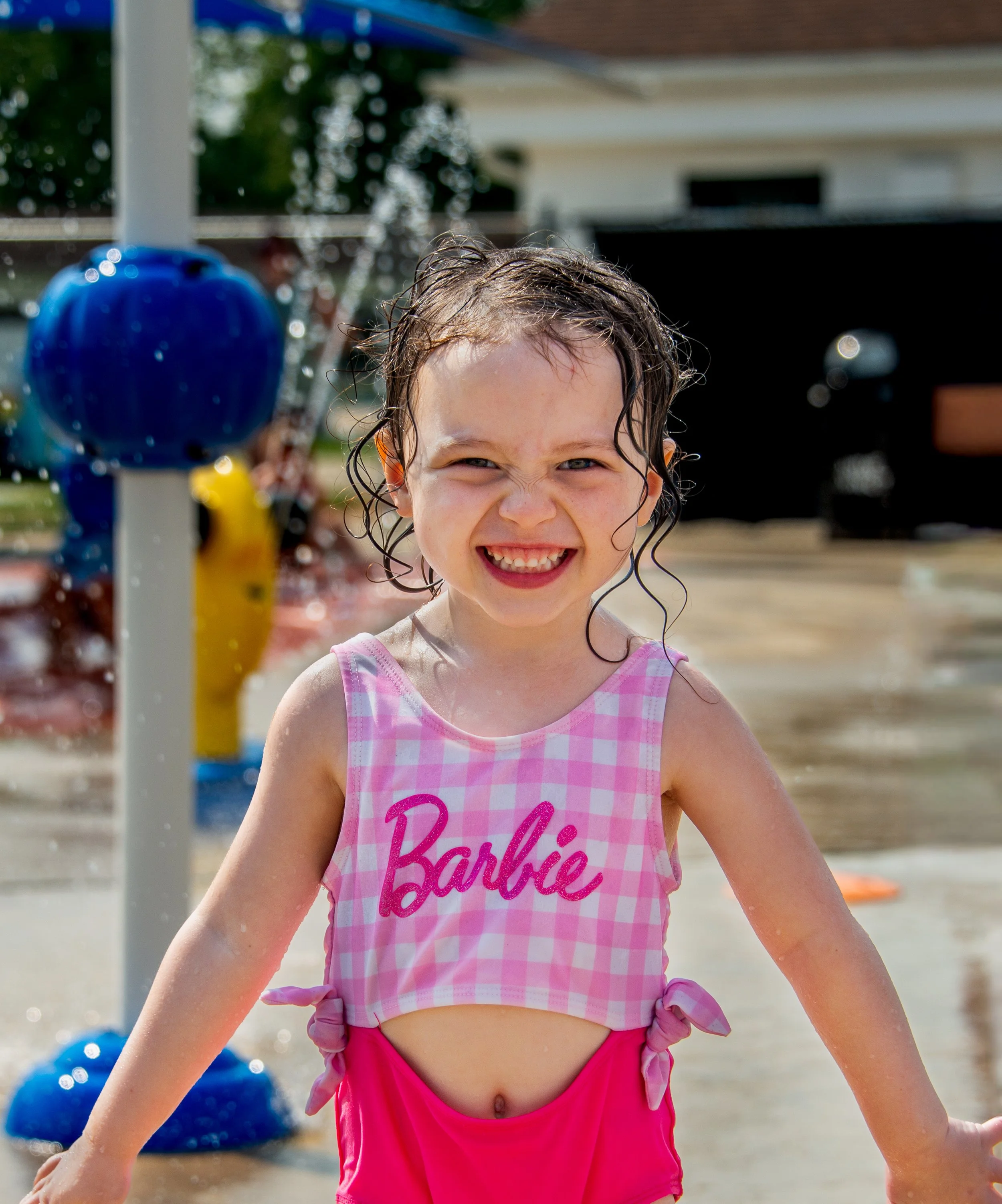 Smiling young girl at a splash park wearing a pink checkered top with 'Barbie' written on it and pink shorts, water splashing around her.