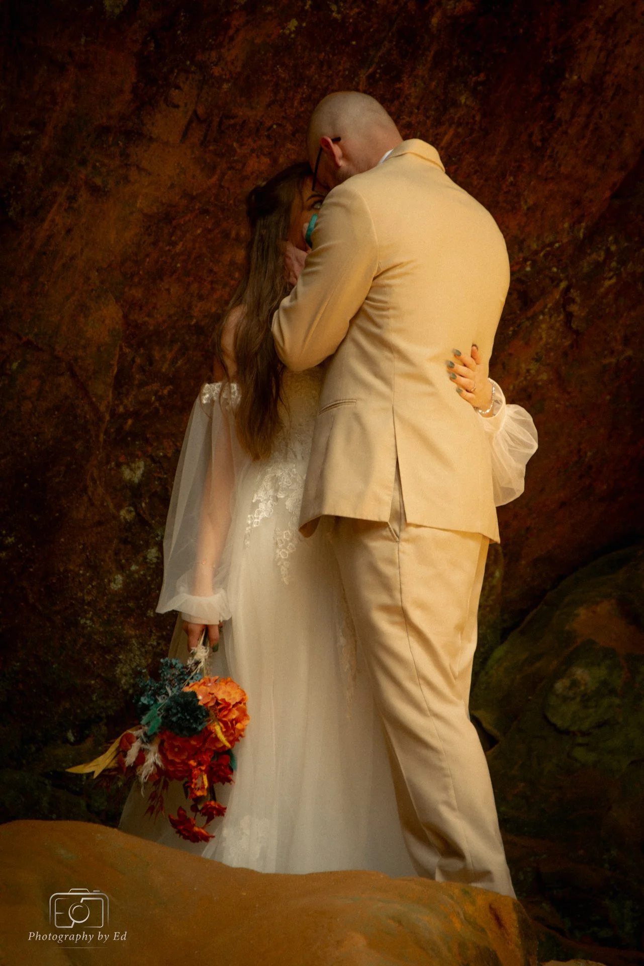 A man and a woman, dressed in formal wedding attire, sharing an intimate moment. The woman holds a bouquet, and they are surrounded by rocky, natural scenery.