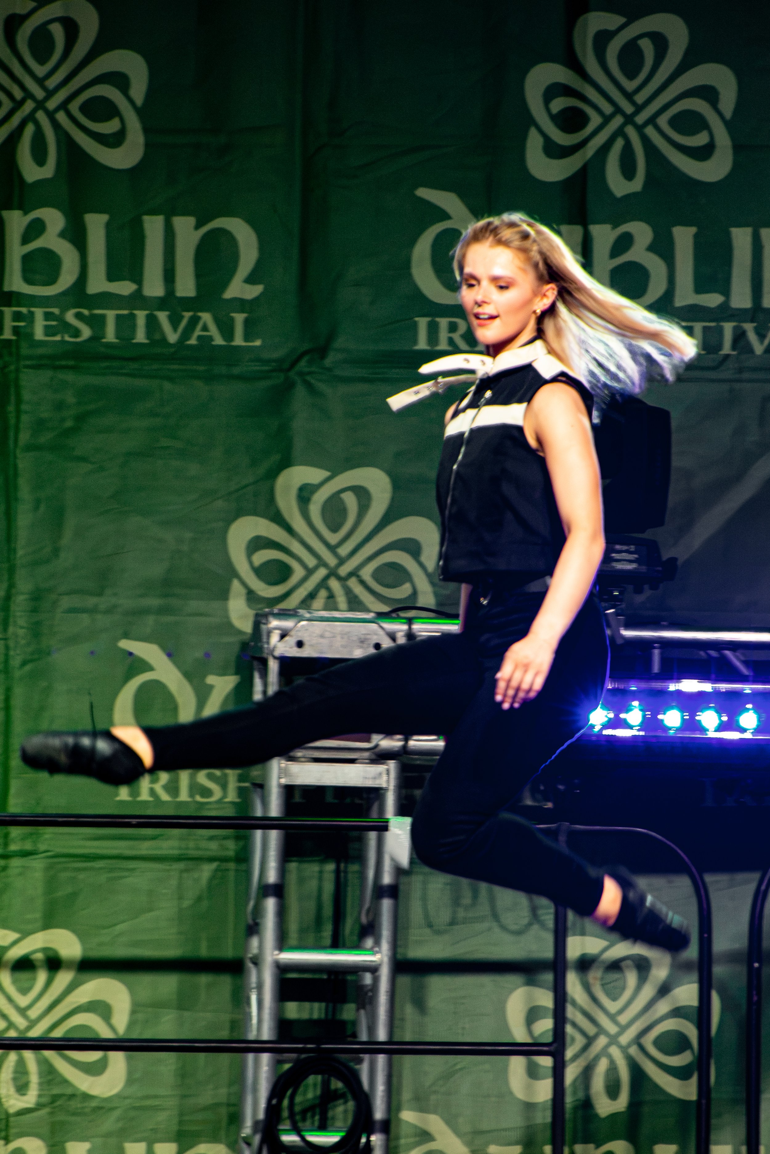 A woman performing a dance move or pose on stage at the Ireland Irish Festival, with a green backdrop displaying the festival's logo and name.