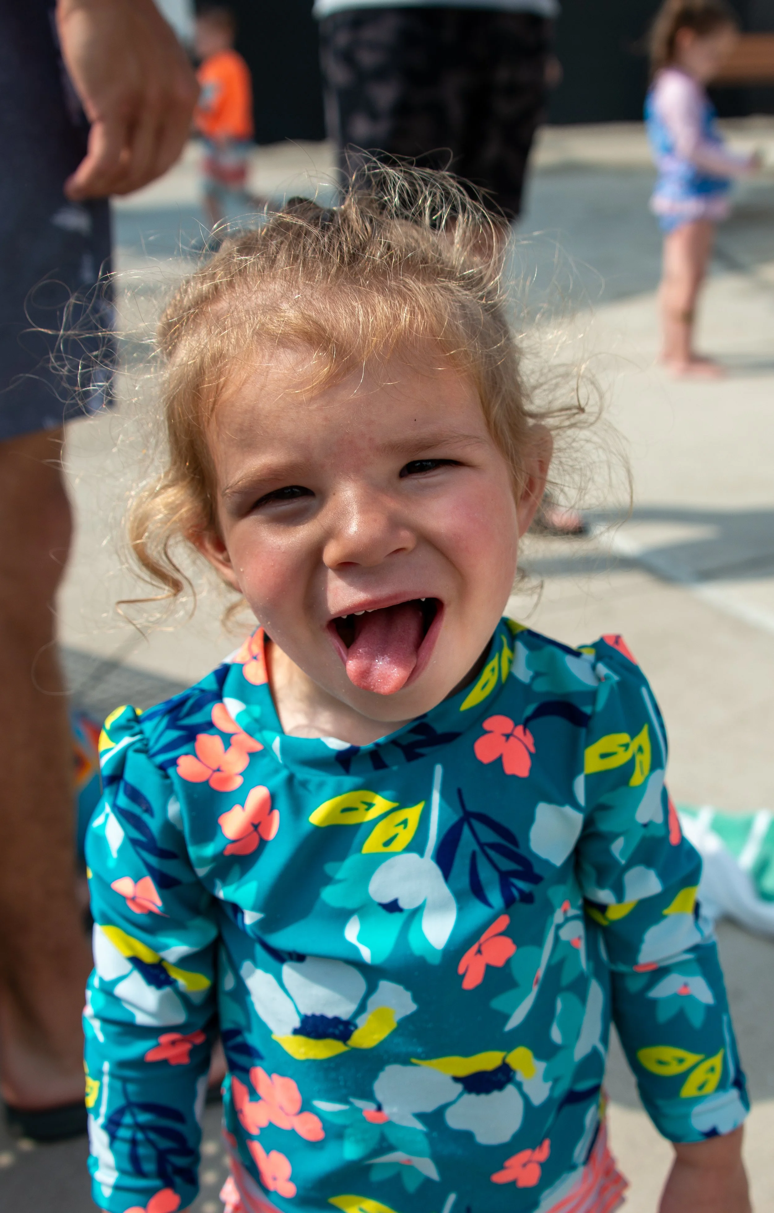 A young child with curly blonde hair sticking out their tongue, wearing a colorful Hawaiian shirt with flowers and leaves, outdoors on a sunny day.