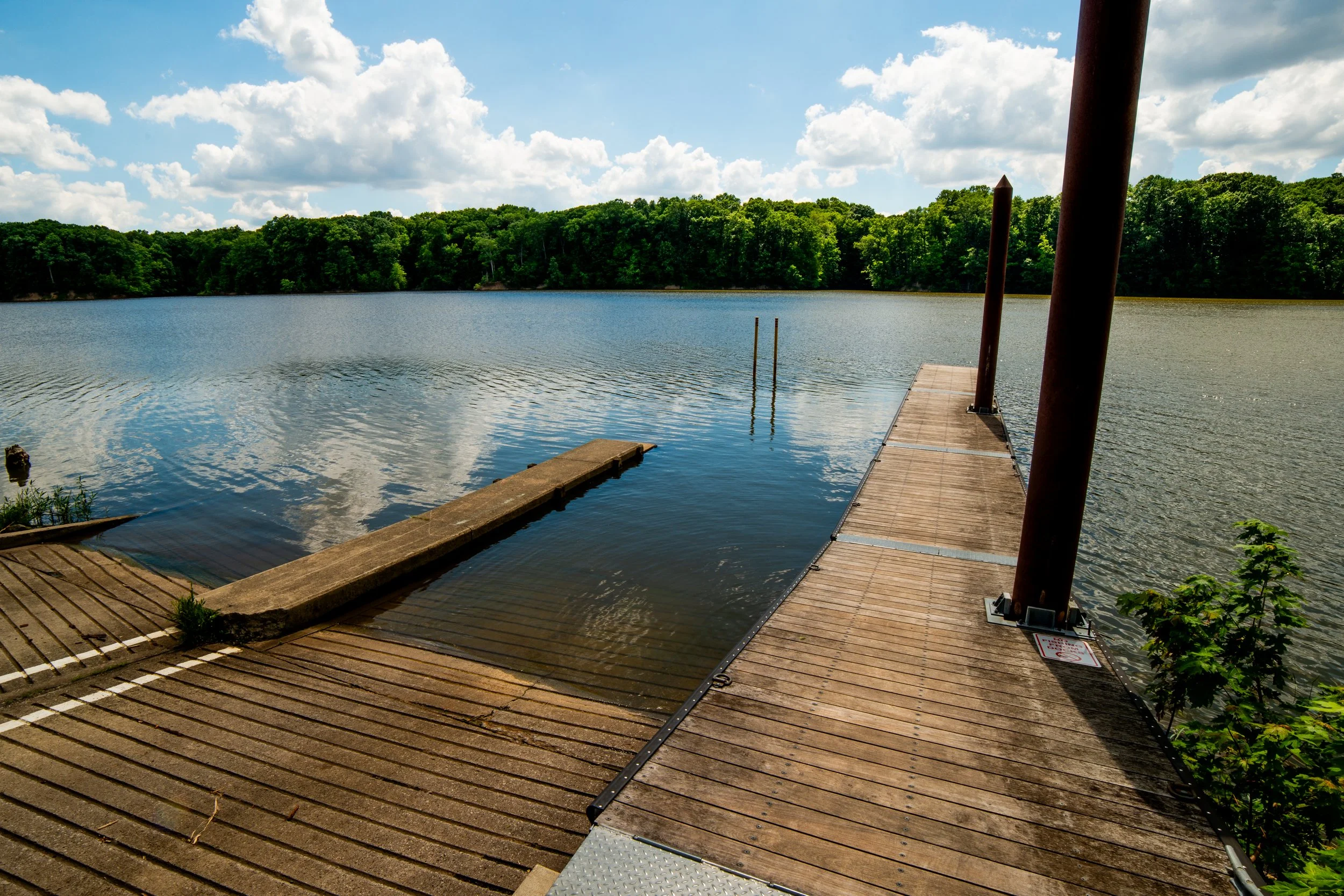 A wooden dock extends into a calm, blue lake surrounded by lush green trees under a partly cloudy sky.