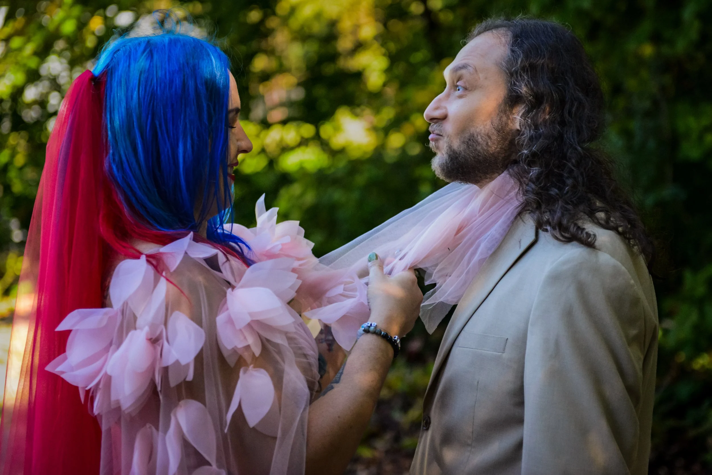 A woman with blue and pink hair wearing a pink dress gently holding the face of a man with long curly hair and a beard, wearing a beige suit, in a garden setting.