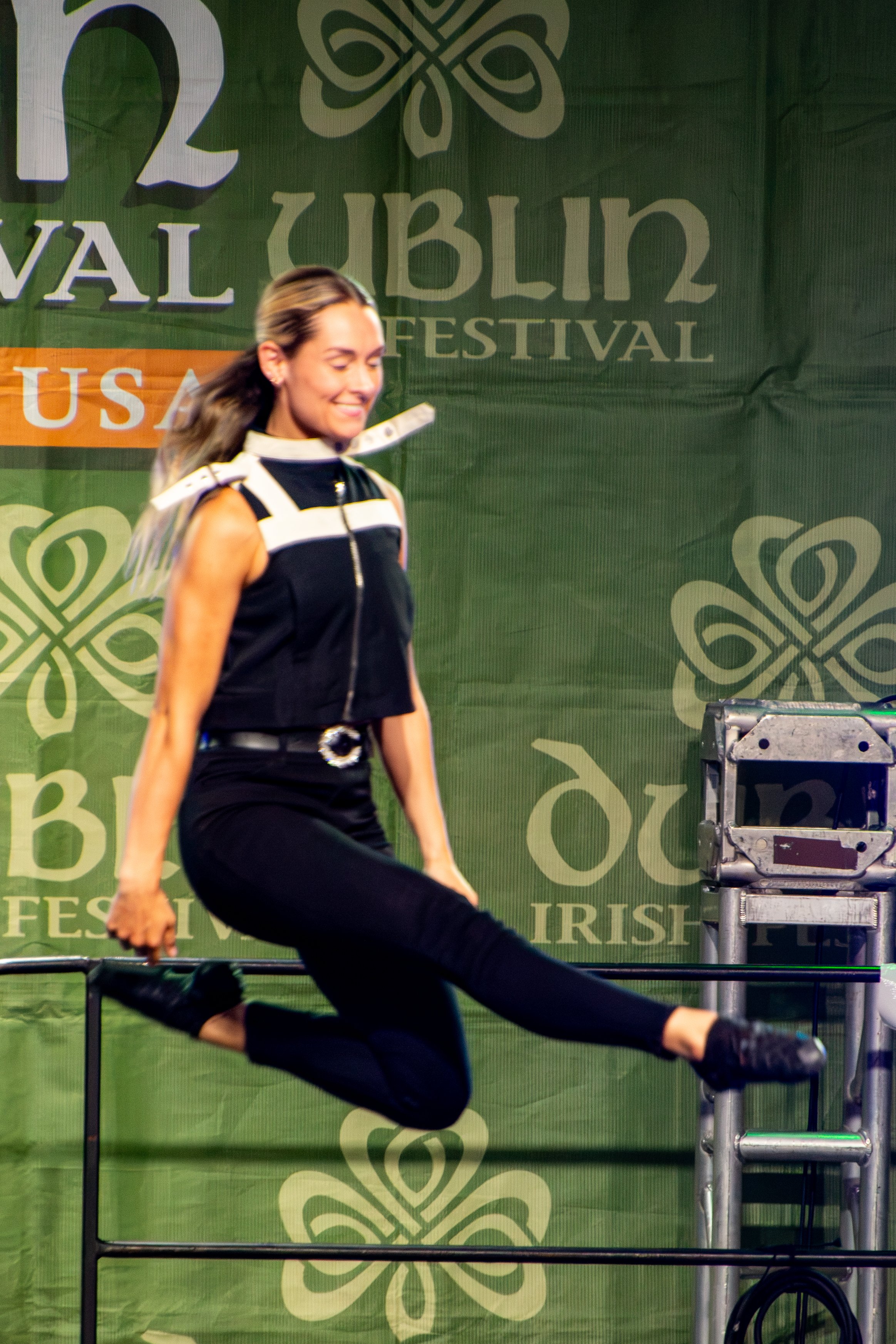 A woman with long hair smiling and performing a dance move on stage at the Irish festival, with a large green banner in the background that reads '11th Irish Festival USA' and decorative shamrock patterns.