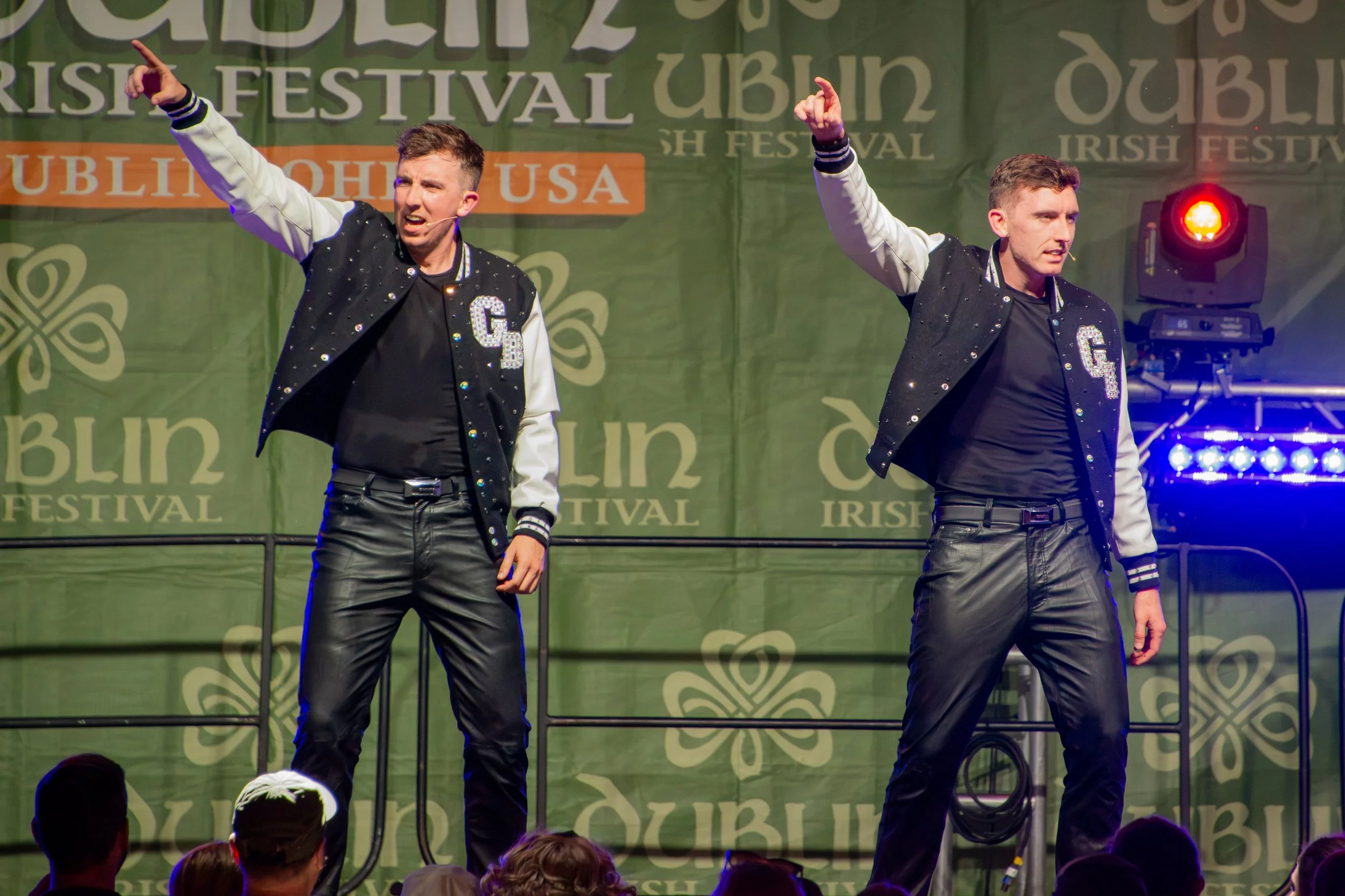 Two male performers on stage at an Irish festival, wearing black and white varsity jackets, singing and dancing with their right arms raised. Audience members visible at the bottom of the image.