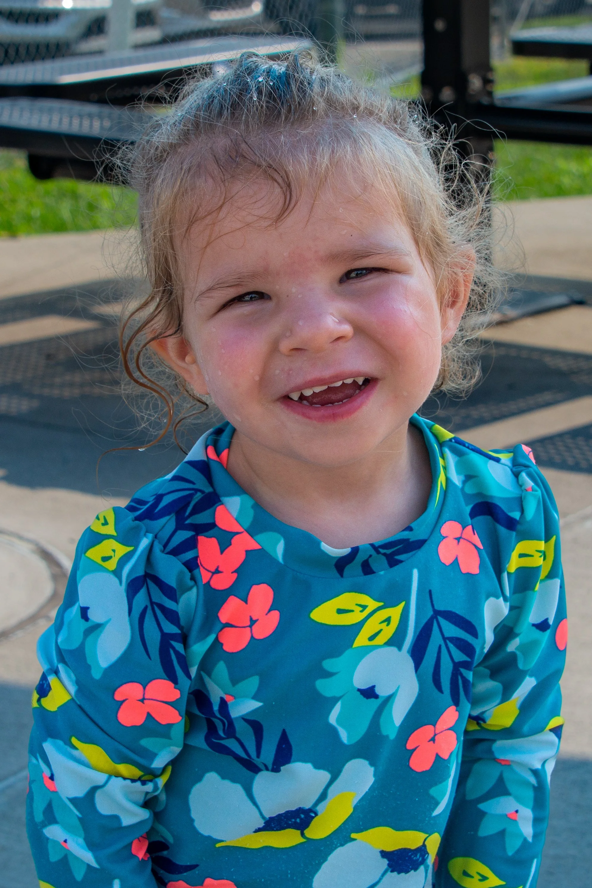 A young girl with wet hair wearing a brightly colored floral swimsuit, smiling and standing outdoors near a park bench.