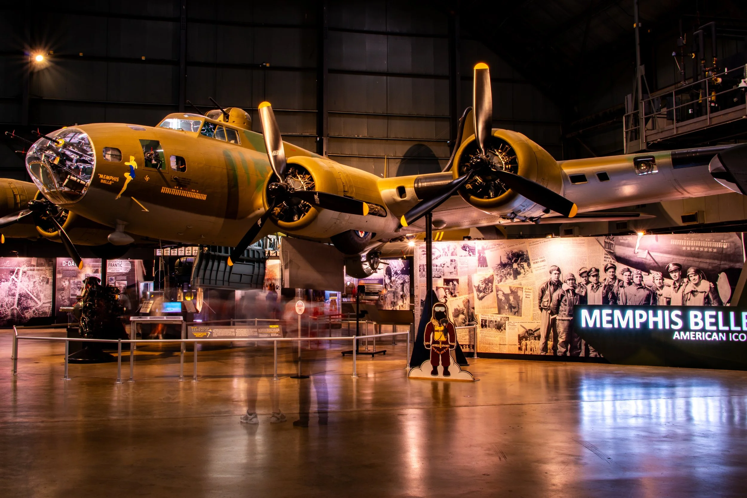 A vintage wreckage of a Memphis Belle American bomber aircraft inside a museum, with informational displays and historical photos on the wall.