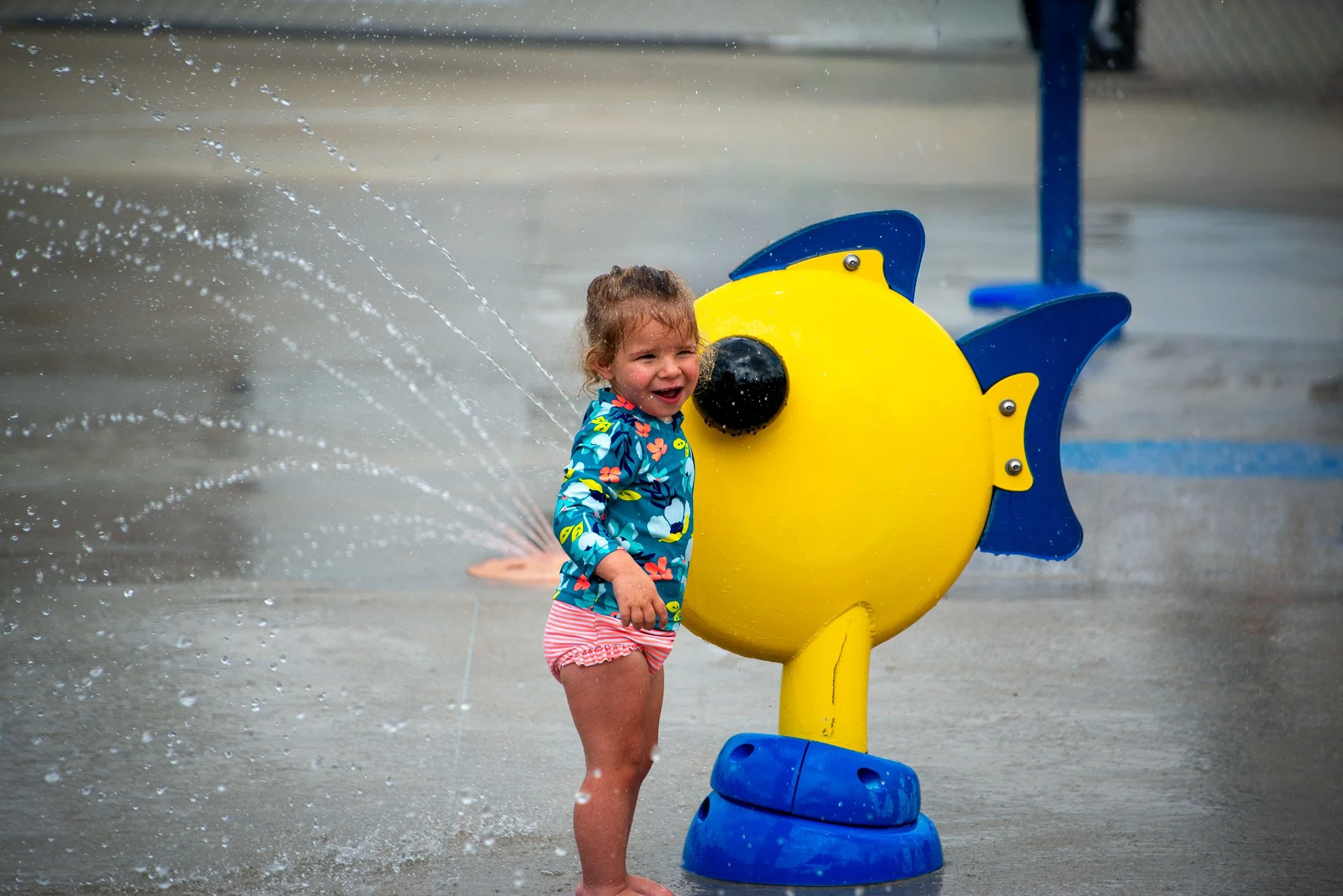 Young girl with wet hair playing at a splash pad with a blue and yellow fish-shaped water fountain, smiling and enjoying the water spray.