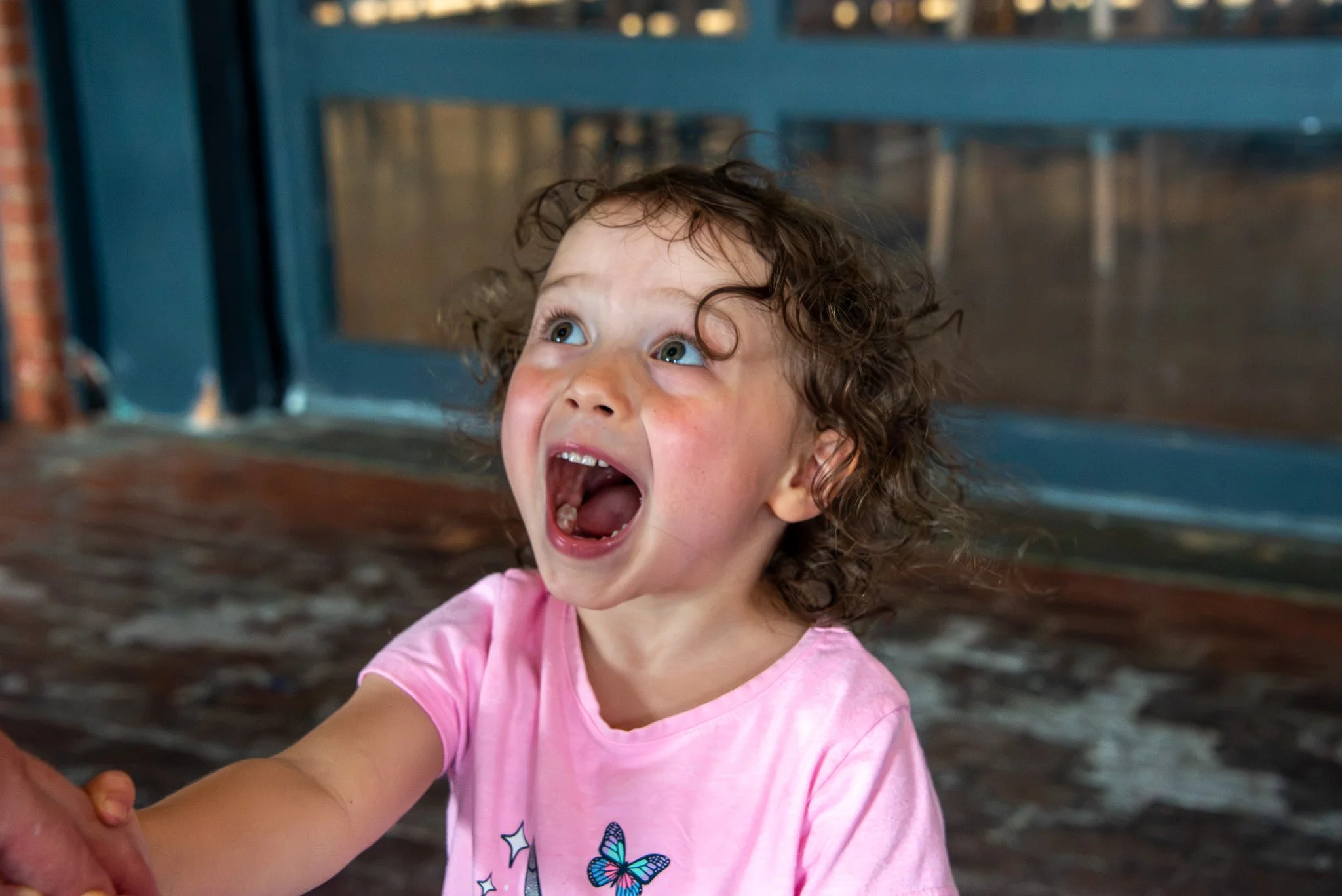 A young girl with curly hair and blue eyes is smiling with her mouth open while holding hands with someone. She is wearing a pink shirt with butterfly and star designs. The background shows a blue structure with a glass window.