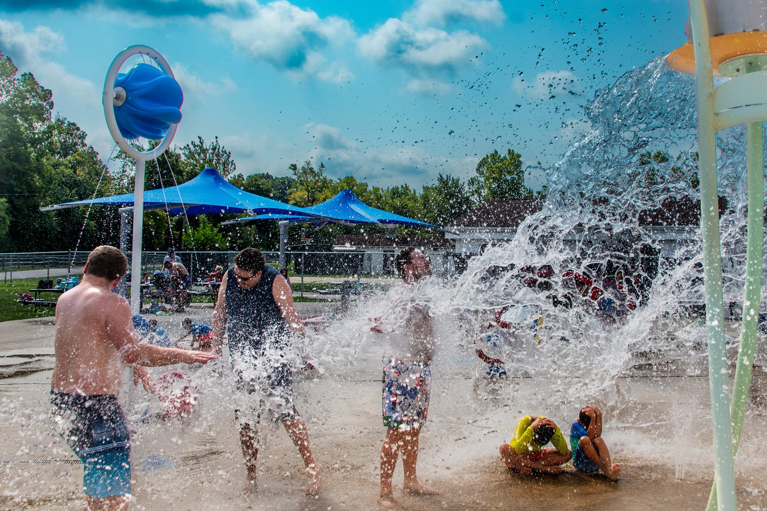 Children and adults playing in a splash pad with water sprays and fountains, some sitting and some standing, under a partly cloudy sky with trees in the background.