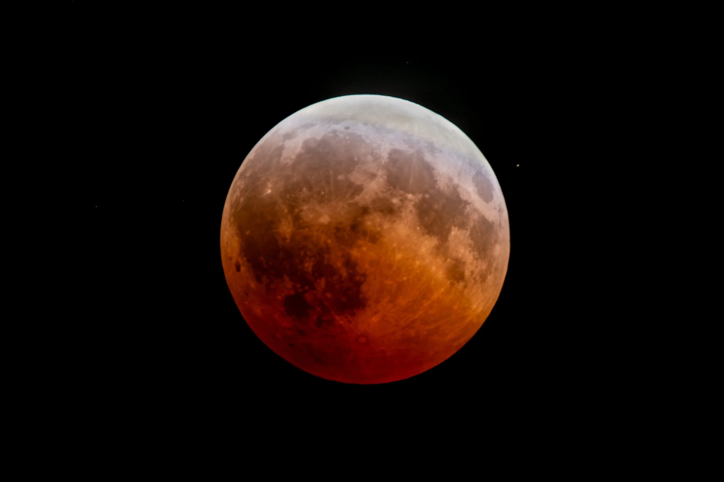 A detailed photograph of a lunar eclipse showing the moon with a reddish hue and some craters on its surface, set against a black night sky with a few distant stars.