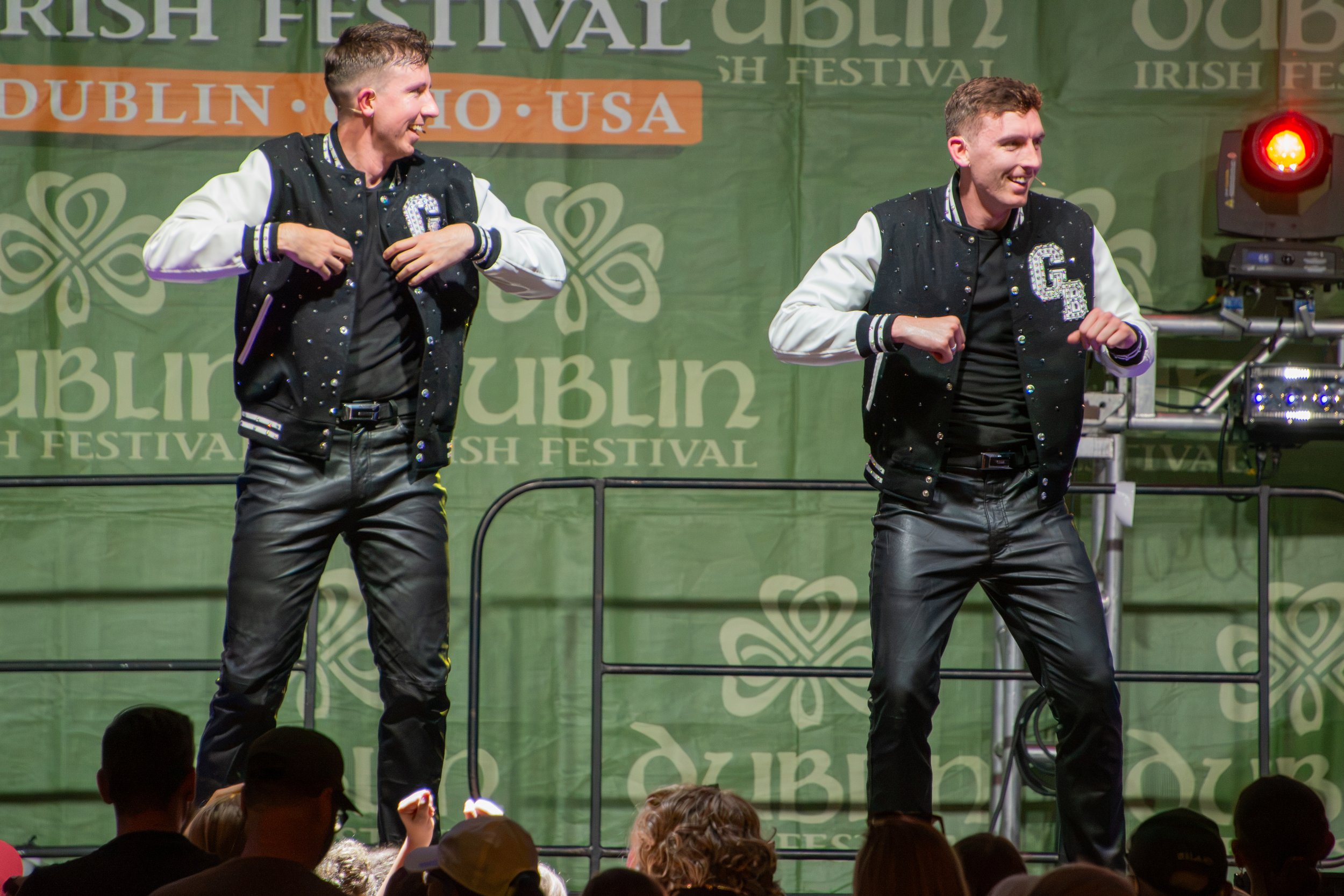 Two male dancers performing on stage at the Irish Festival in Dublin, Ohio, with a green Irish-themed backdrop labeled 'Irish Festival' and 'Dublin, Ohio, USA.' They are wearing black and white jackets with letter patches, and are dancing in front of
