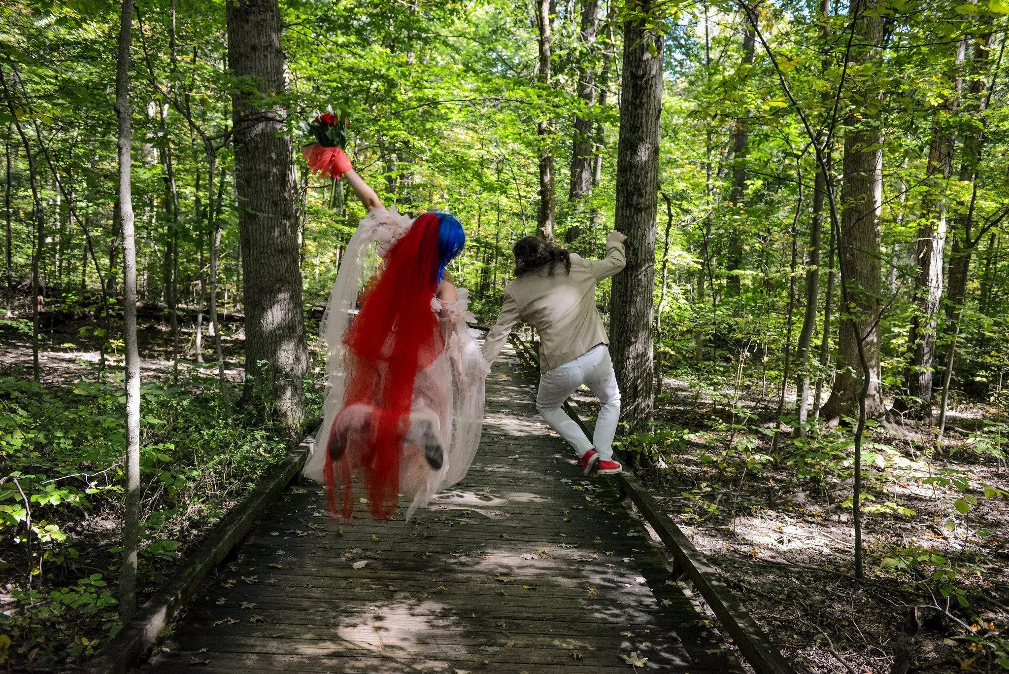 A person with rainbow-colored hair in a dress, holding a bouquet of flowers, running on a wooden trail in a dense green forest, assisted by another person in a beige jacket and white pants.