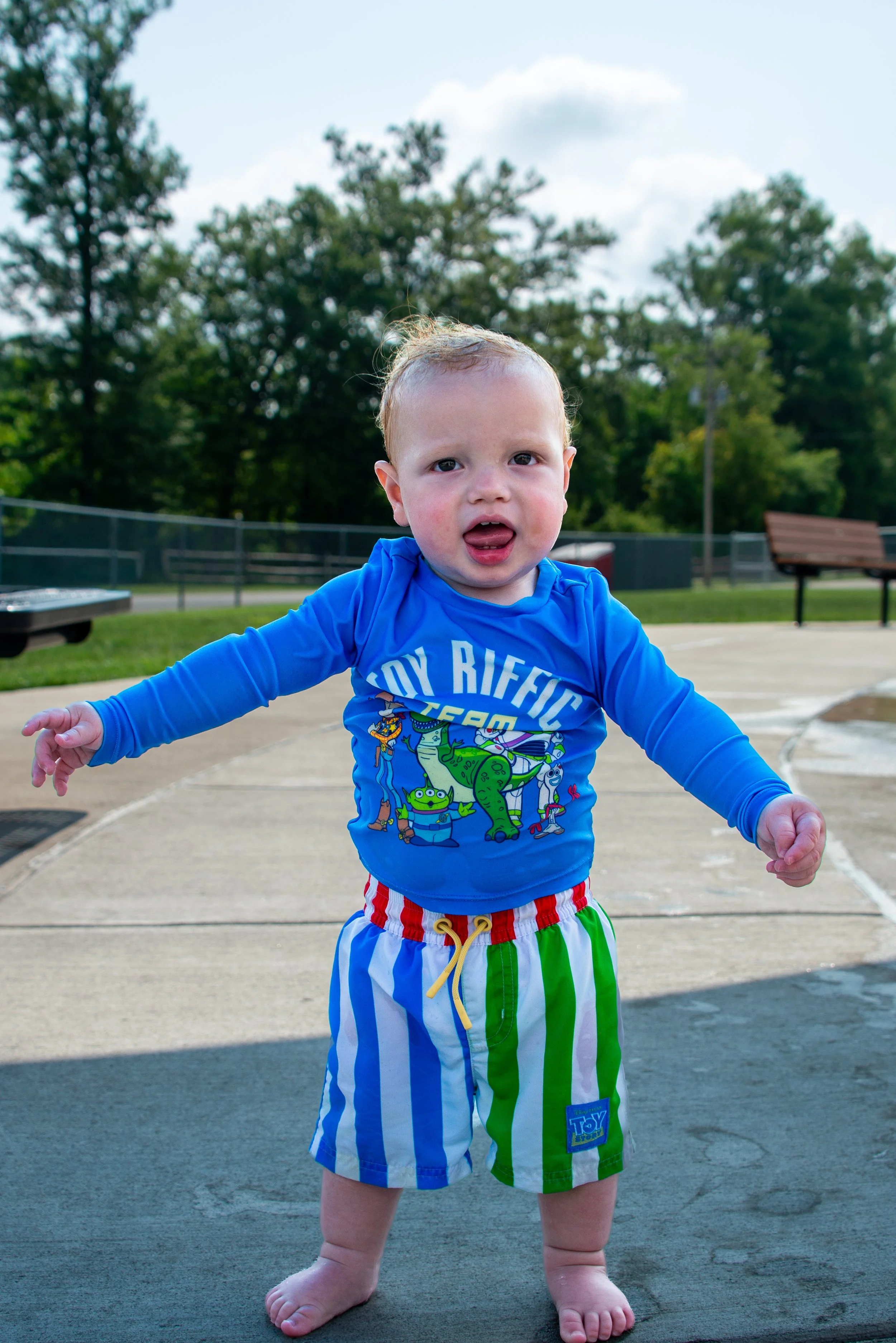 Young boy outdoors in a park with trees and benches in the background, wearing a blue Toy Story shirt and colorful striped shorts, barefoot, with arms outstretched and tongue slightly out.