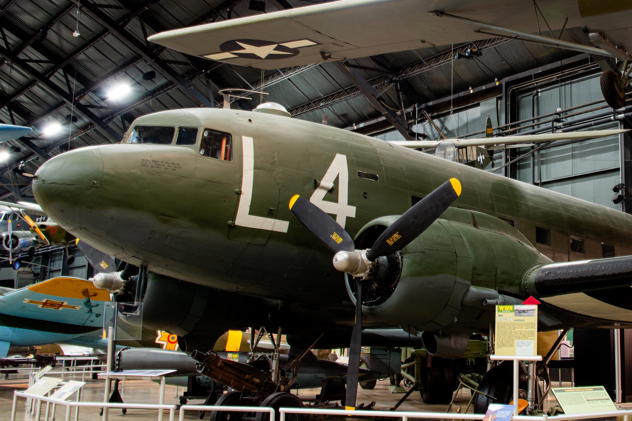 A military aircraft on display inside a museum, with a large propeller and various markings, including a white star and a number 4, underneath a ceiling with metal beams and lighting.