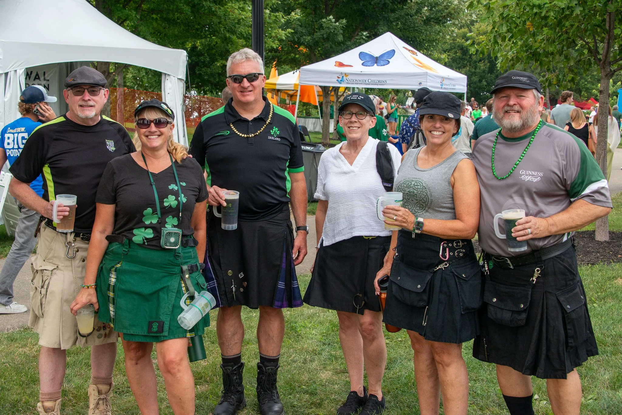 A group of seven people smiling and holding drinks at an outdoor event with tents and trees in the background, some wearing casual and some in Irish-themed attire, including a kilt and shamrock accessories.