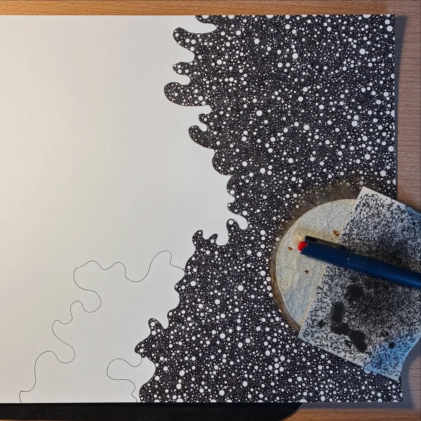 Studio overhead view of process work by contemporary minimalist Abdushakur, showing dense black ink stippling and organic accumulation growing from the top edge on archival board, alongside a pen and ink test fragment.
