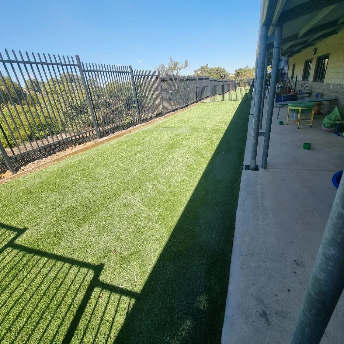 Freshly laid artificial grass on a backyard lawn, with a black metal fence along one side and a concrete patio on the other, under a clear blue sky.