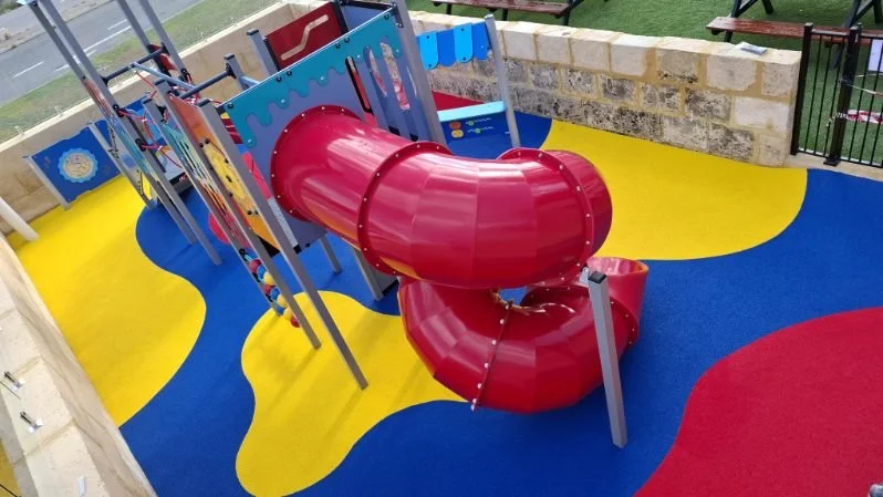 Colorful playground with a red spiral slide, blue and yellow rubber flooring, and a brick wall in the background.
