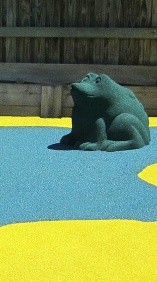 A frog-shaped sculpture on a colorful playground surface with a wooden fence in the background.