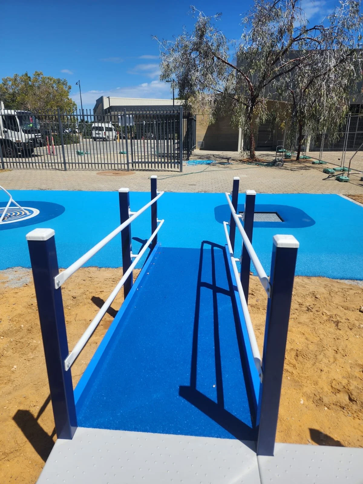 A blue wheelchair accessible ramp leading to a playground area with a blue rubber surface, surrounded by a dirt area. In the background, there are trees, a parking lot with cars, and a building under a clear blue sky.
