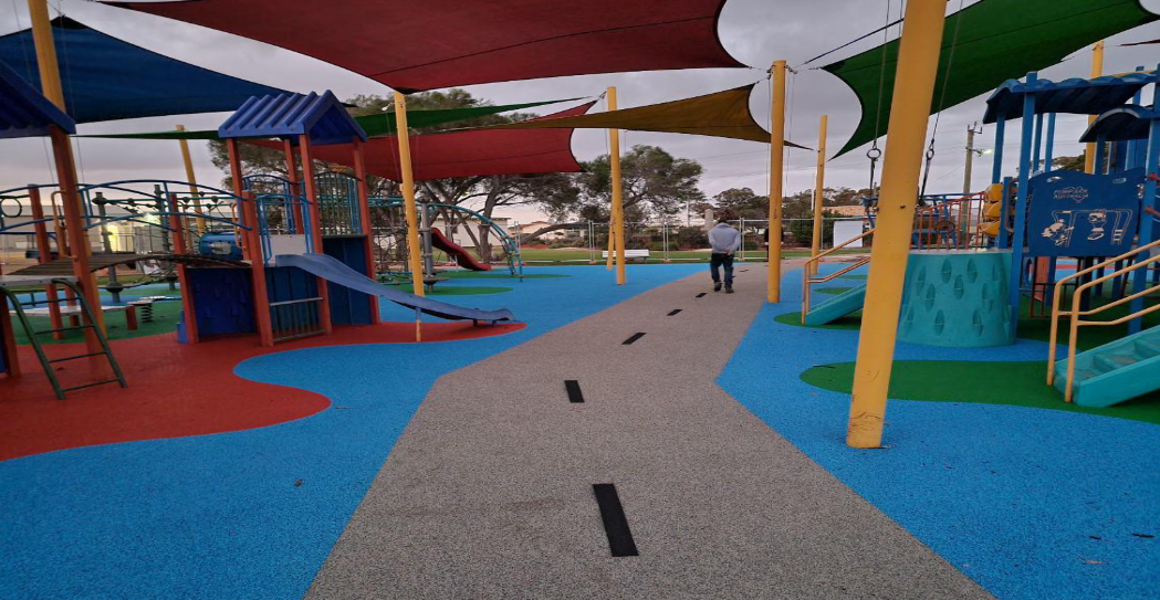 Colorful playground with slides, climbing structures, and shaded areas, featuring a squirrel sculpture on the right side, with a person walking towards the back.