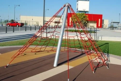 Red and black rope climbing structure on a playground with a multicolored rubber surface, in a park with a sidewalk and a modern building in the background.