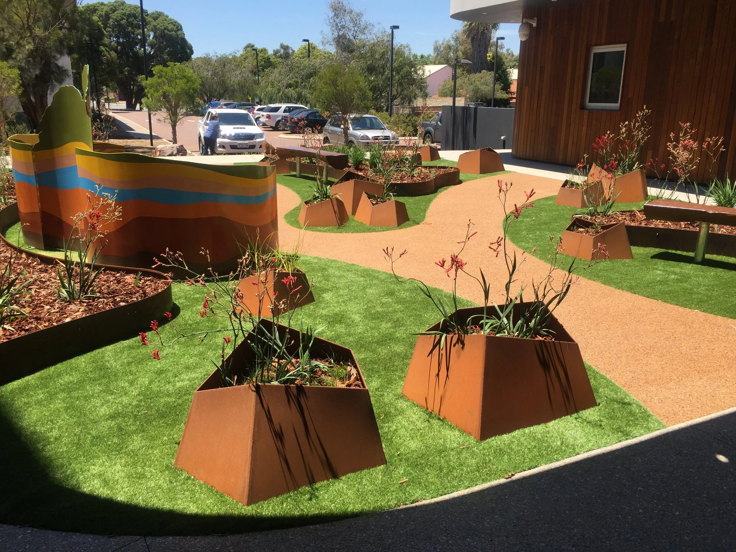 Outdoor landscaped area with a colorful, striped large sculpture, small garden beds with plants, and a cement pathway, with parked cars and trees in the background.