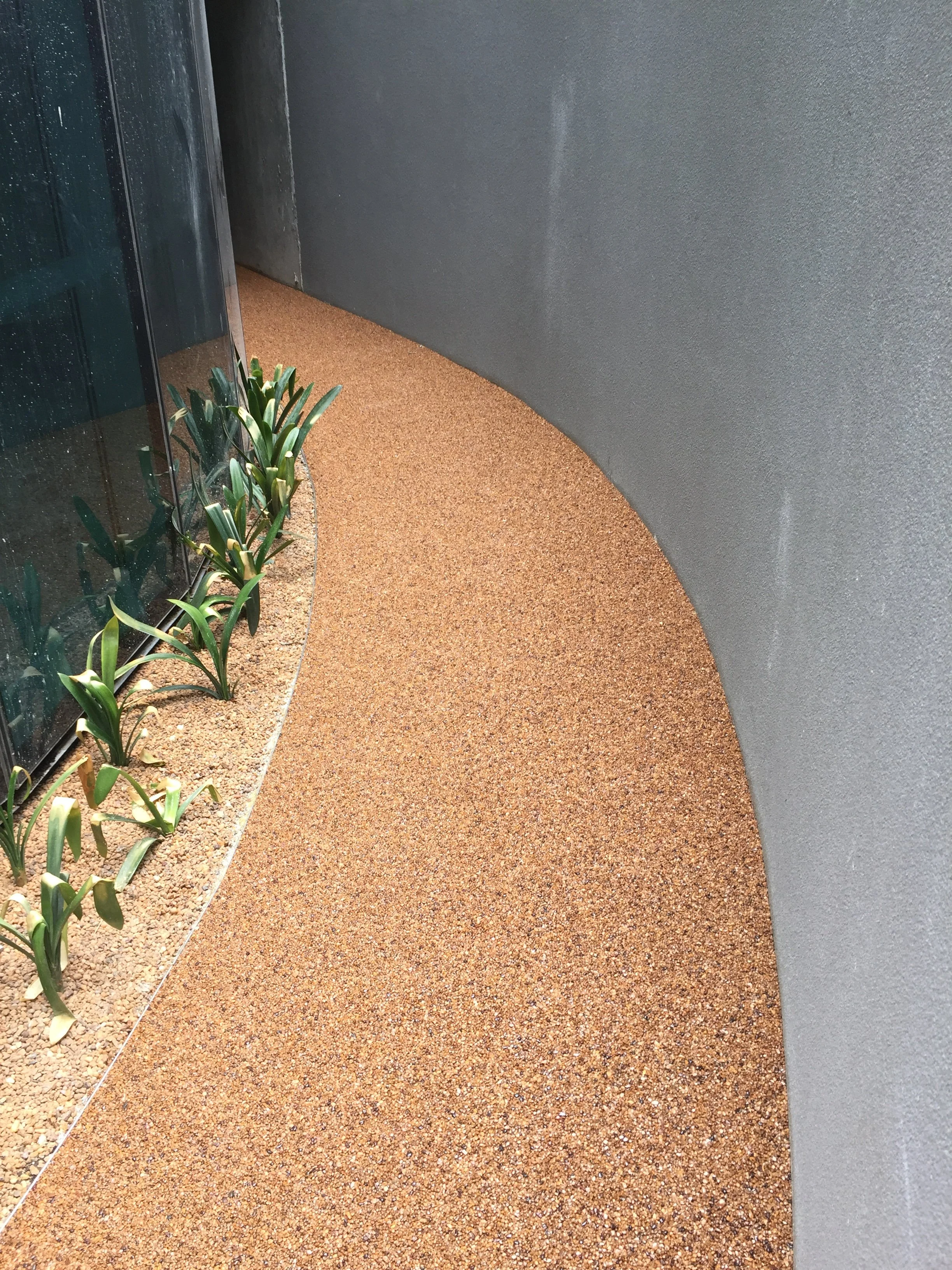 Smooth brown textured pathway alongside a glass wall with green plants.
