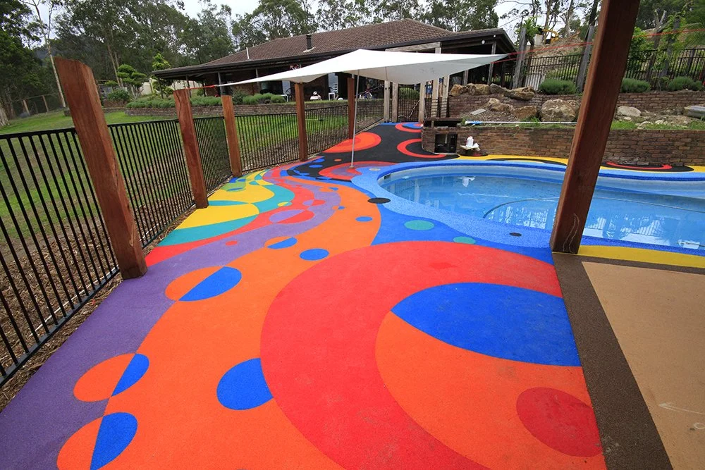 Colorful poolside walkway with a bright, abstract pattern of orange, blue, purple, yellow, and red circles and swirls, enclosed by a black metal fence, with a pool to the right and a shaded seating area in the background.
