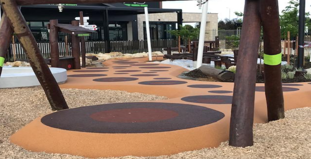 Playground with colorful rubber flooring featuring large circles, trees, and outdoor seating in the background.