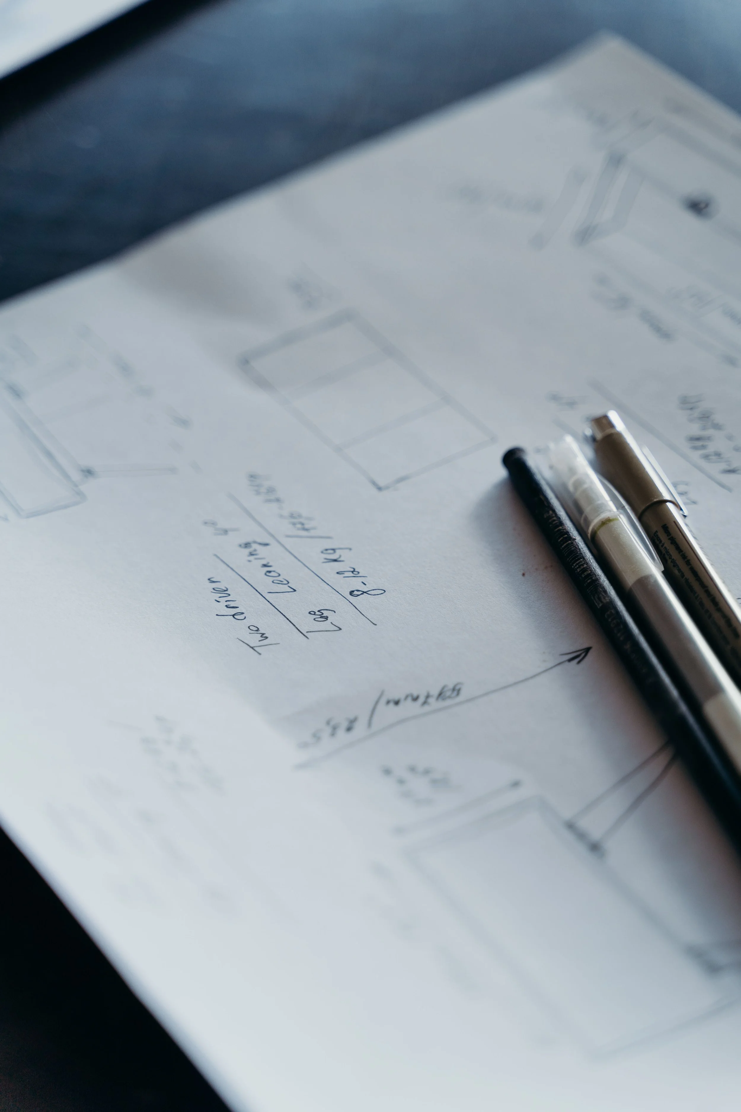 Close-up of handwritten notes and sketches on paper with two pens, on a desk.