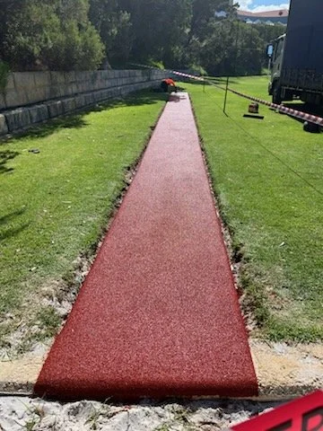 A newly installed red jogging or walking track on a grassy area, with construction equipment and caution tape in the background.