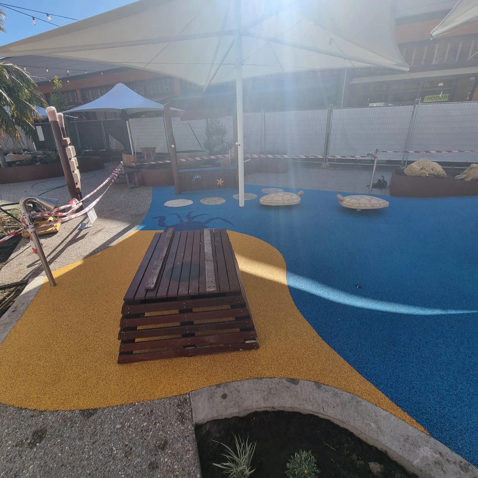 Outdoor playground with a colorful rubber surface, a wooden bench, shade structures, and decorative rocks.