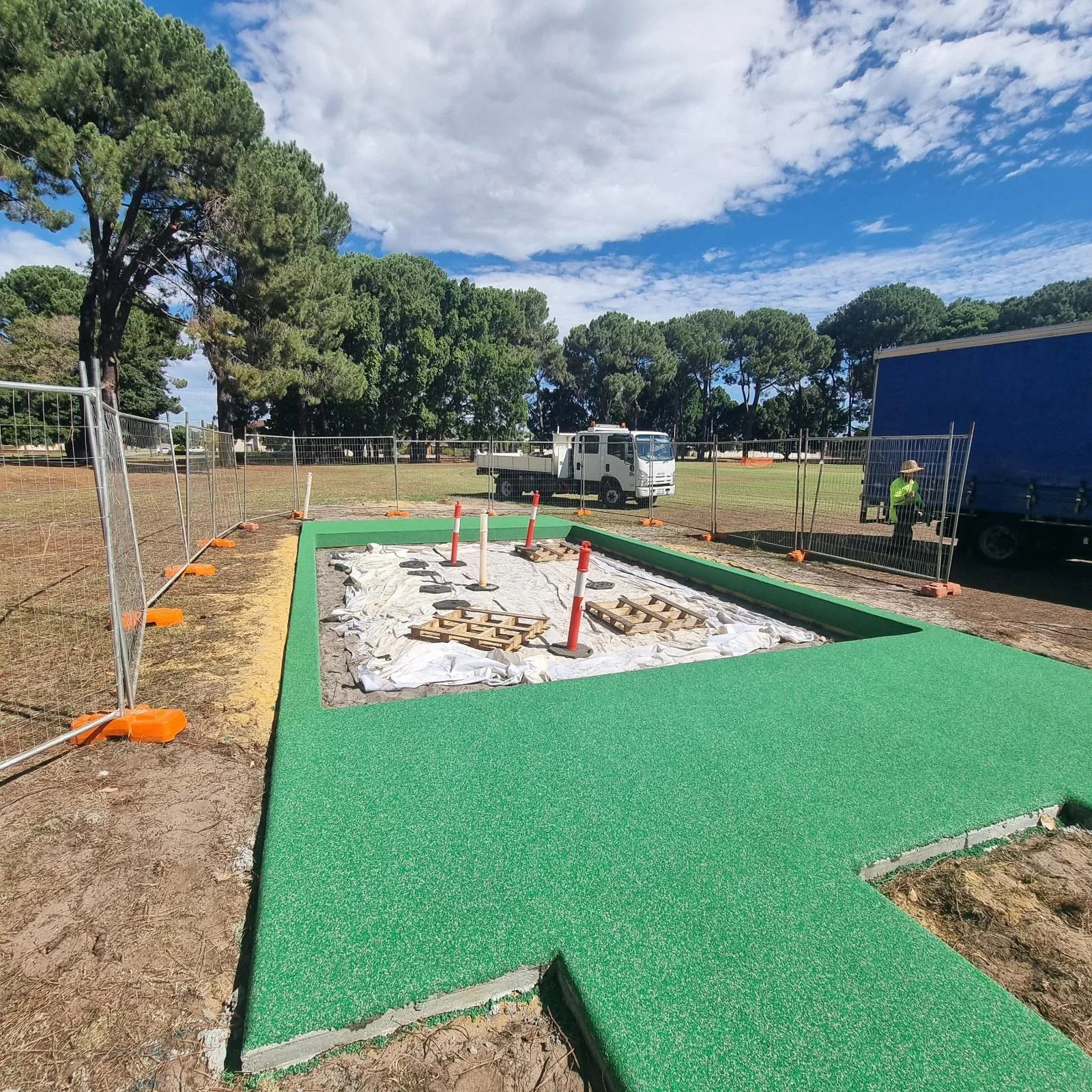 Construction site with a green artificial turf border, dirt inside, and orange safety cones, with trees and trucks in the background under a partly cloudy sky.