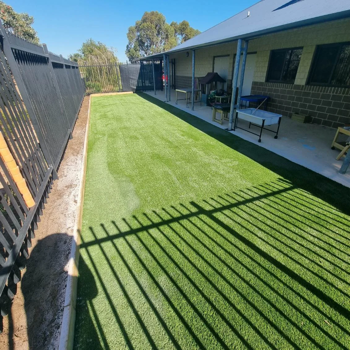 Backyard with freshly laid artificial grass, surrounded by a black metal fence and patio with outdoor furniture, under a clear blue sky.