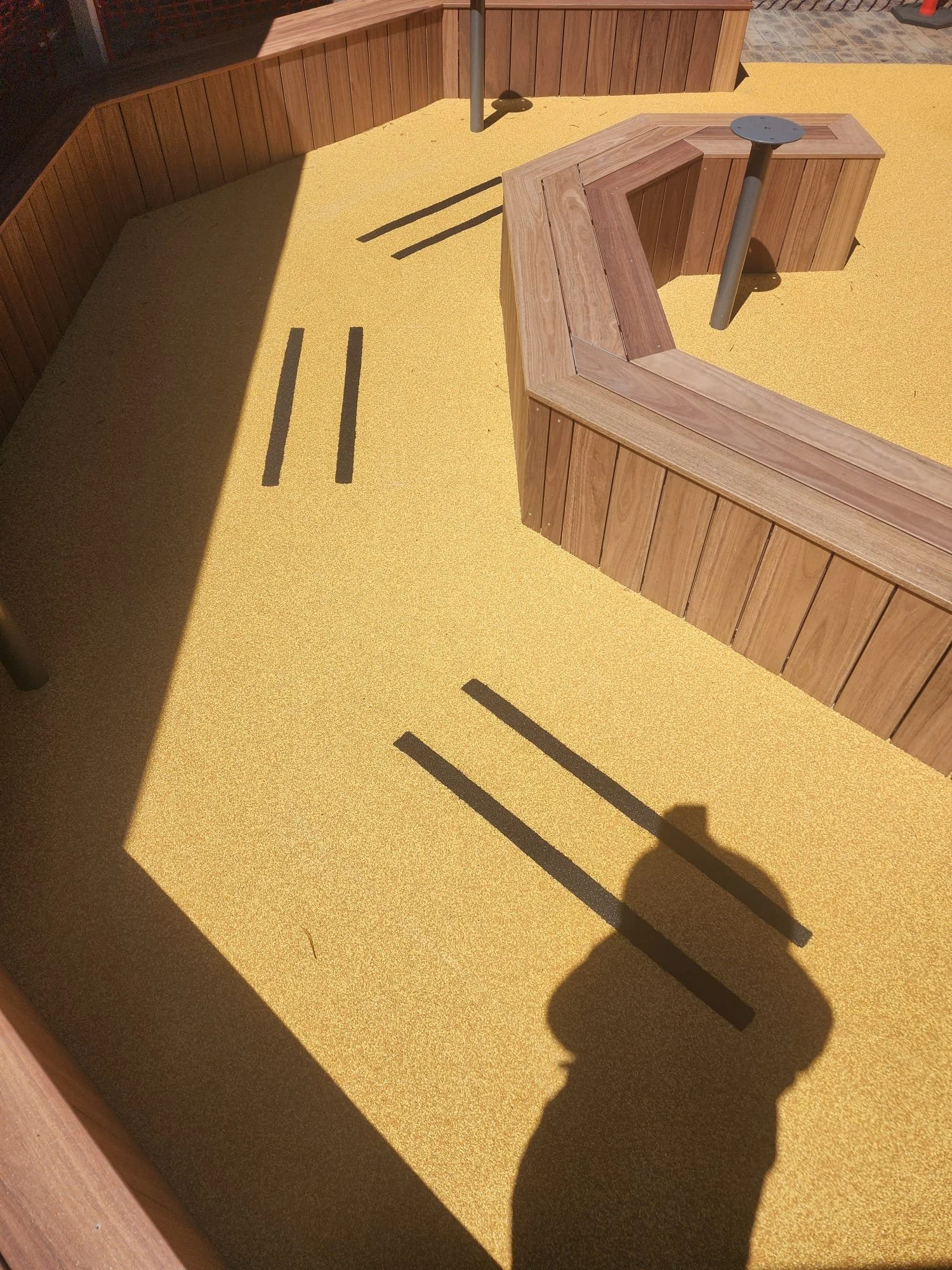 Empty outdoor space with a bright yellow ground covering, bordered by wooden panels, featuring two wooden benches and metal poles casting shadows.