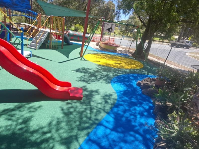 Colorful playground with a red slide, kids' play structure, swings, sandbox, and shaded area, surrounded by trees and a fence.