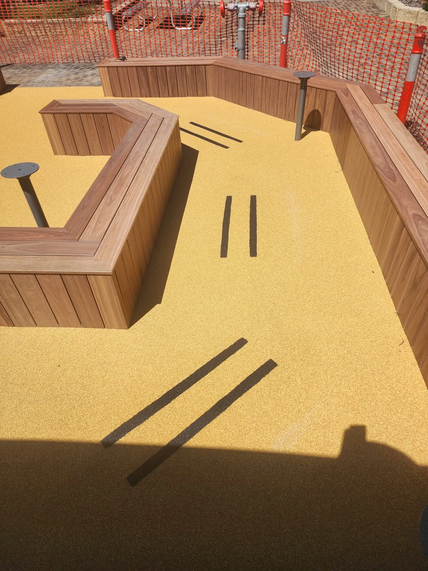 View of playground area with curved wooden benches, black lines on yellow ground surface, construction fencing, and construction equipment in background.