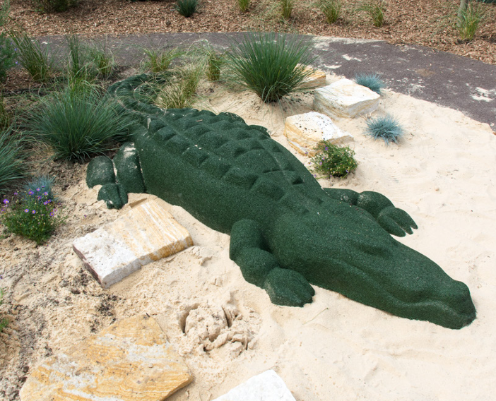 Artificial crocodile sculpture in desert landscape with rocks and plants.