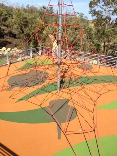 Colorful playground climbing structure with orange and green rubberized surface, surrounded by trees and fencing.