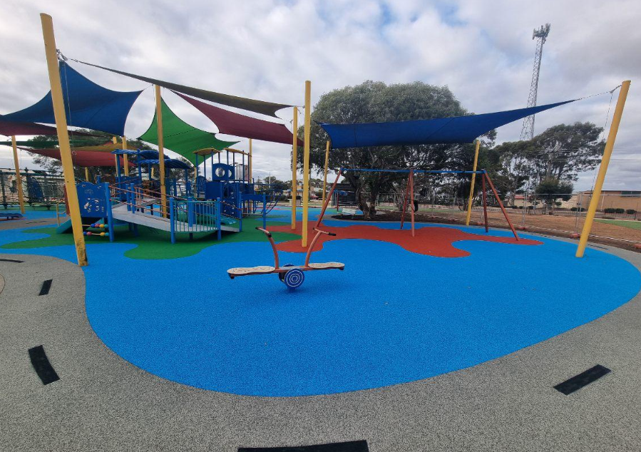 Empty playground with colorful rubber flooring, shade sails, a slide structure, and a seesaw.