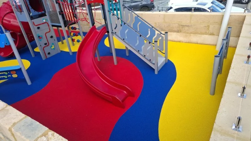 Colorful playground area with red slide, blue and yellow rubber flooring, and climbing structures.