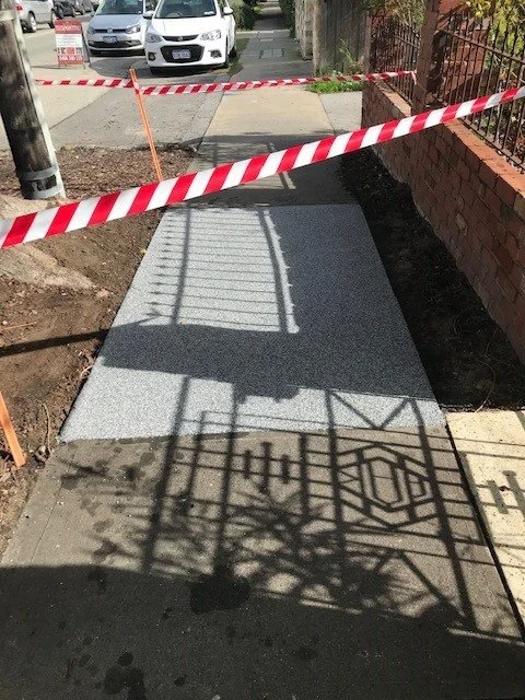 Sidewalk under construction blocked by red and white caution tape, with shadows of a fence and gate cast on the sidewalk.