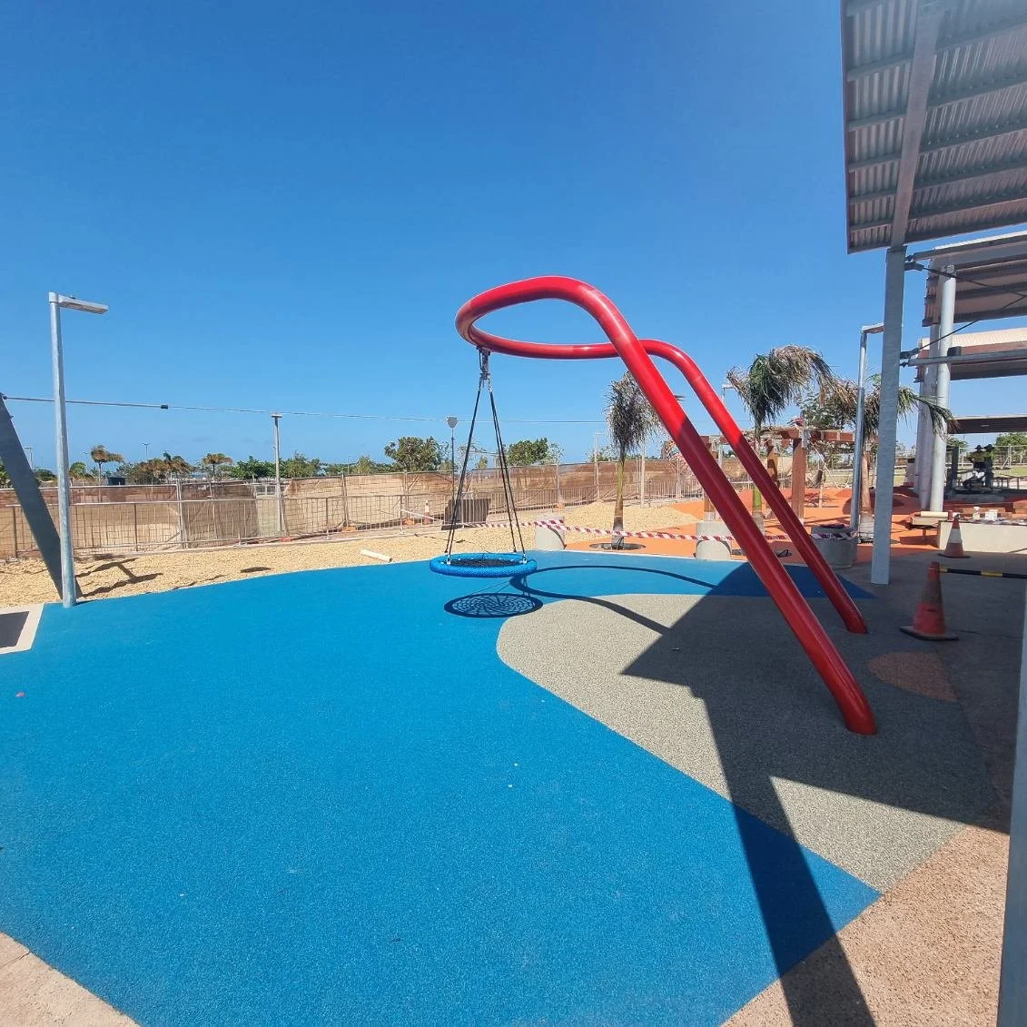 Empty playground with a red slide, a swing, and colorful rubber flooring under a clear blue sky.