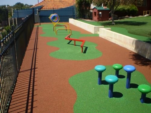 Outdoor playground area with various colorful play equipment on a rubberized surface, enclosed by a fence.