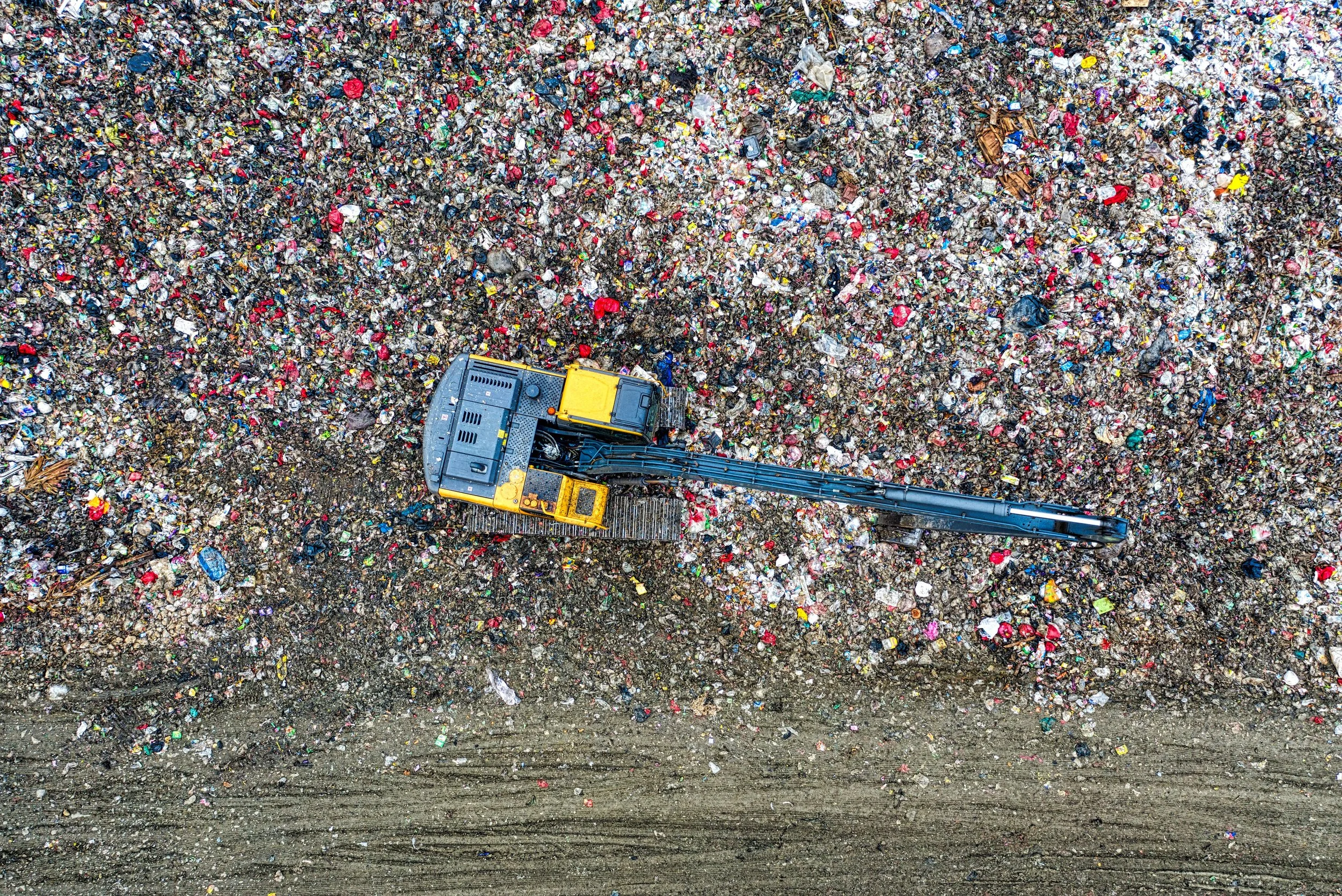 An aerial view of a large landfill filled with scattered plastic, paper, and other waste. A yellow and black excavator is at the bottom center, moving through the trash.