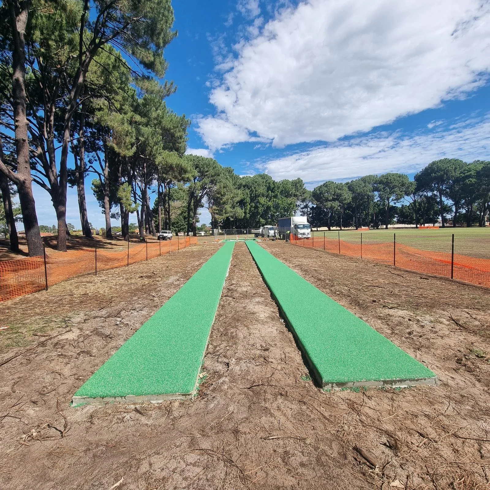 Two green mats laid on the ground in a field, with orange fencing on either side, and trees in the background under a partly cloudy sky.